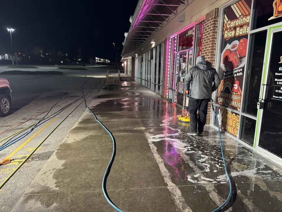 Technician cleaning a shopping center sidewalk at night with a surface cleaner and hoses